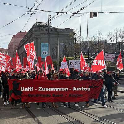 Menschen demonstrieren mit roten Fahnen und einem Banner «Uniti – Zusammen – Juntos» auf einer Strasse in der Stadt.