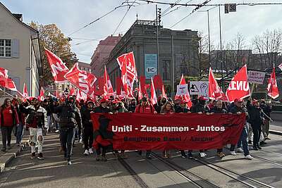 Menschen demonstrieren mit roten Fahnen und einem Banner «Uniti – Zusammen – Juntos» auf einer Strasse in der Stadt.