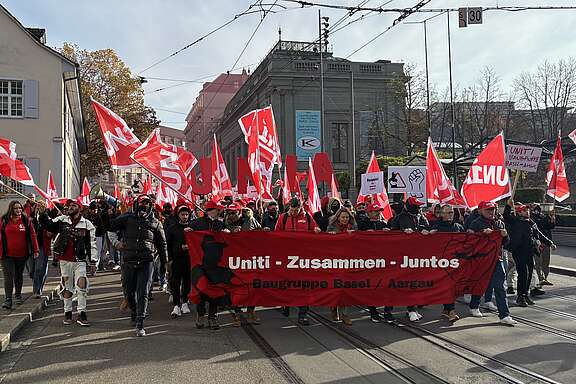 Menschen demonstrieren mit roten Fahnen und einem Banner «Uniti – Zusammen – Juntos» auf einer Strasse in der Stadt.