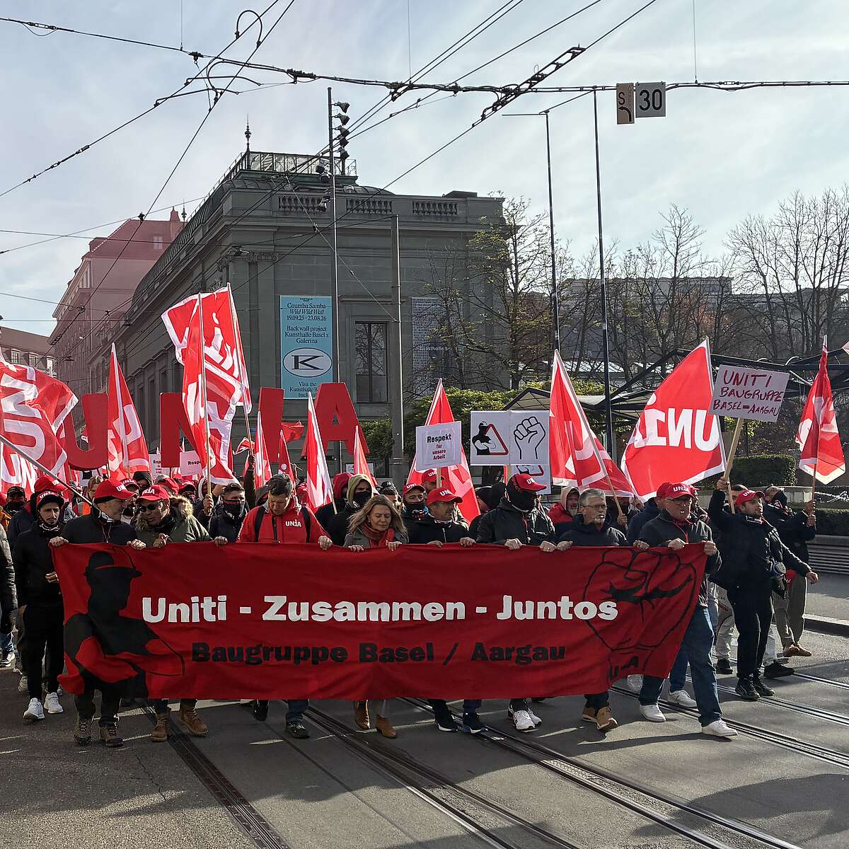 Menschen demonstrieren mit roten Fahnen und einem Banner «Uniti – Zusammen – Juntos» auf einer Strasse in der Stadt.