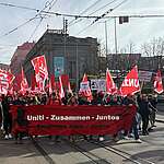 Menschen demonstrieren mit roten Fahnen und einem Banner «Uniti – Zusammen – Juntos» auf einer Strasse in der Stadt.