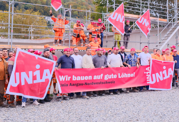 Eine Gruppe von Bauarbeitern posiert auf einer Baustelle mit einem grossen roten Transparent der Gewerkschaft Unia, auf dem steht: «Wir bauen auf gute Löhne! Unia Aargau-Nordwestschweiz».